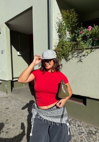 Hall värviline müts, millel on rhinestones, punane sobiv T-särk, mustad laiad püksid ja oliivroheline kott. Taustal on roheline sein ja taimed.