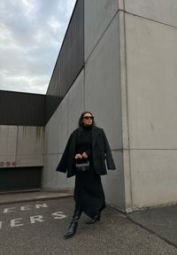 Črn oversized plašč preko črne obleke, v kombinaciji z črnimi škornji do kolen. Drži majhen črn torbici. Siva betonska ozadje.