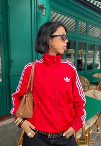 Moteris raudoname Adidas treniruočių paltuose ir akinių nuo saulės stovi su rankomis kišenėse prie kavinės, kurios sienos žali, o kėdės pintos.