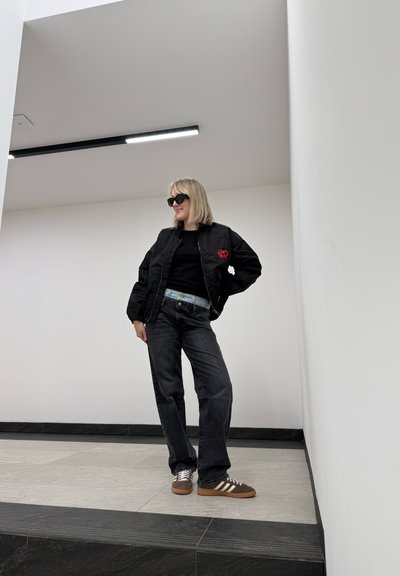 Chaqueta bomber acolchada negra con bordado rojo, camiseta negra, jeans rectos grises y zapatillas Adidas marrones con rayas blancas.