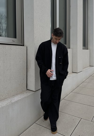Hombre con chaqueta negra, camisa blanca y pantalones negros caminando por un camino de concreto junto a un edificio moderno con grandes ventanas.