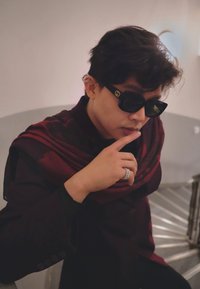 Personne portant des lunettes de soleil noires avec un logo doré, un tissu drapé bordeaux, des vêtements sombres et une bague argentée, posant avec un doigt sur les lèvres.