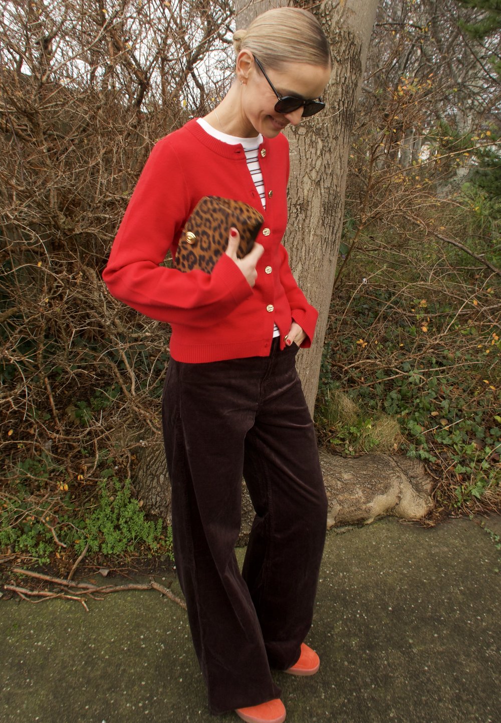 Cardigan rouge avec boutons dorés, haut rayé, pantalon large en velours côtelé marron, et chaussures orange. Tenant une pochette à imprimé léopard.