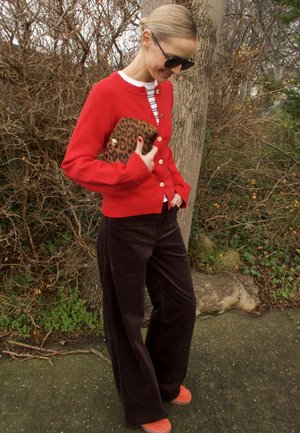 Cárdigan rojo con botones dorados, camiseta de rayas, pantalones anchos de pana marrón y zapatos naranjas. Sosteniendo un bolso de mano con estampado de leopardo.