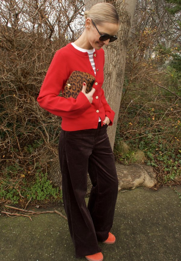 Cardigan roșu cu nasturi aurii, top cu dungi, pantaloni maro tip largi din catifea cu rețea și pantofi portocalii. Ține un clutch cu imprimeu leopard.