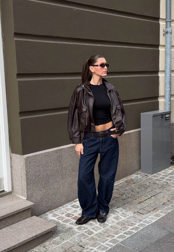 Una mujer con gafas de sol, una chaqueta de cuero oscura, un top negro y vaqueros de pierna ancha está de pie en una calle adoquinada junto a un edificio.