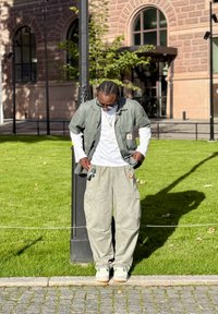 Mężczyzna z zaplecionymi warkoczami, noszący okulary przeciwsłoneczne, luźną warstwową zielono-białą koszulę i beżowe spodnie typu cargo, stojący na brukowanej drodze obok trawy i budynku.