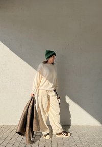 Suéter de malha bege, calças creme e ténis com padrão de leopardo. Segurando uma camada exterior castanha, usando um gorro verde contra um fundo claro.