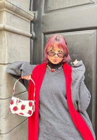 Borsa bianca con motivo di fragole rosse, maglione grigio, cardigan rosso e dolcevita nero. I capelli sono rosa, incorniciati da occhiali da sole scuri.