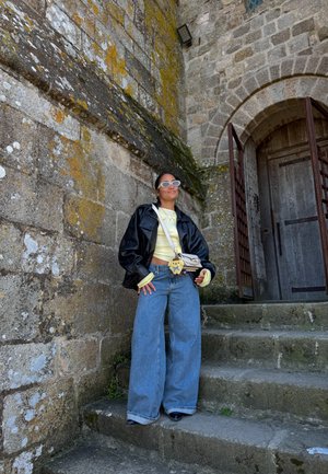 Persona que lleva una chaqueta de cuero negra, camiseta amarilla, vaqueros anchos azules y gafas de sol blancas, de pie en unos escalones de piedra junto a una pared de piedra musgosa y una puerta arqueada de madera.