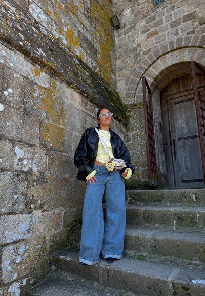 Persona que lleva una chaqueta de cuero negra, camiseta amarilla, vaqueros anchos azules y gafas de sol blancas, de pie en unos escalones de piedra junto a una pared de piedra musgosa y una puerta arqueada de madera.