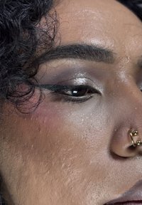 Nærbilde av et ansikt med mørkt, krøllete hår, sminke som inkluderer sølvøyenskygge og sveipet eyeliner, samt en gull septum-piercing.