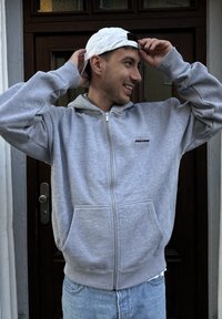 Grå hettejakke med glidelås og en frontlomme samt en liten svart logo. Bært sammen med en hvit baseballcaps, mot en bakgrunn av en tre-dør.