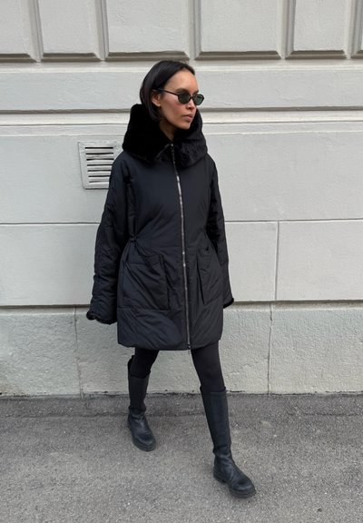Mujer con abrigo acolchado negro con cuello grande, leggings negros, botas y gafas de sol caminando junto a una pared de piedra clara.