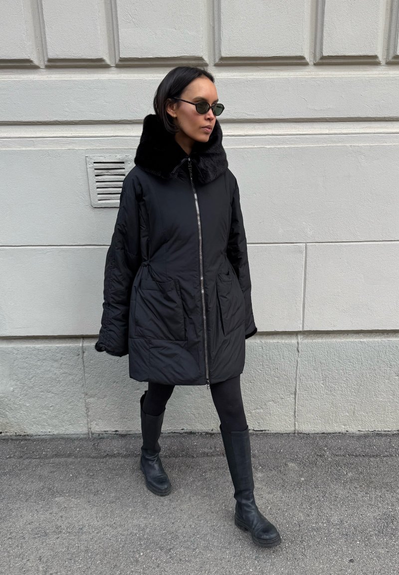 Femme portant un manteau matelassé noir à grand col, des leggings noirs, des bottes et des lunettes de soleil, marchant le long d'un mur en pierre claire.