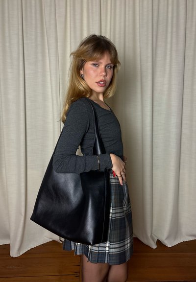 Bolso tote de cuero negro con textura suave, forma curva y correa para el hombro, combinado con una blusa gris y una falda de cuadros.