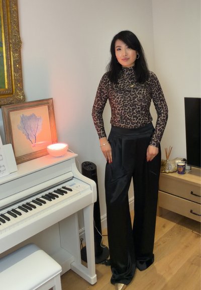 Mujer con cabello negro que lleva un top de estampado de leopardo y pantalones negros, de pie junto a un piano blanco con una obra de arte enmarcada y una lámpara encendida.