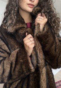 Abrigo de piel sintética marrón con un cuello amplio, textura suave y mangas largas, sostenido por manos que muestran anillos. Presenta sutiles rayas oscuras.