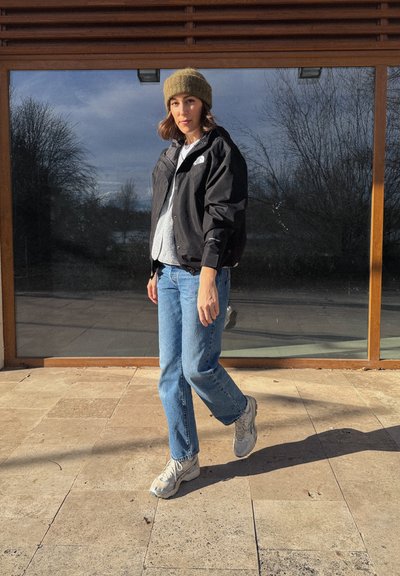 Mujer con gorro de punto marrón, chaqueta negra, suéter gris, vaqueros azules y zapatillas blancas, de pie sobre baldosas de piedra frente a una gran ventana.