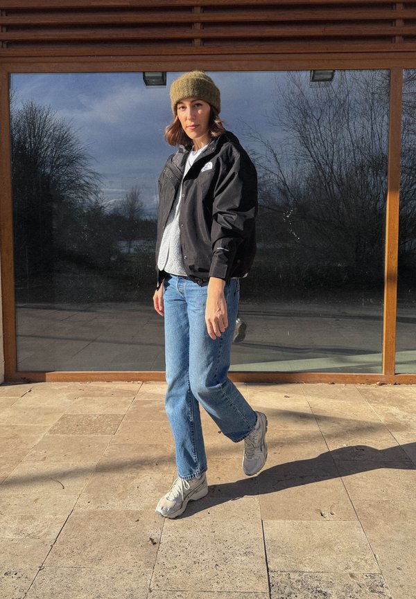 Frau mit brauner Strickmütze, schwarzer Jacke, grauem Pullover, blauer Jeans und weißen Sneakers, die auf Steinfliesen vor einem großen Fenster im Freien steht.