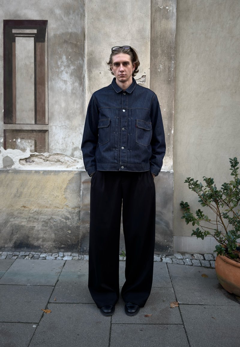 Jachetă din denim cu două buzunare frontale, culoare albastru închis, purtată peste pantaloni negri lejeri și pantofi negri, pe fundalul unui perete texturat.