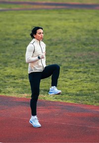 Coureur portant une veste crème clair à fermeture éclair avec des accents noirs et un pantalon noir ajusté, chaussé de chaussures de course bleues, sur une piste rouge.