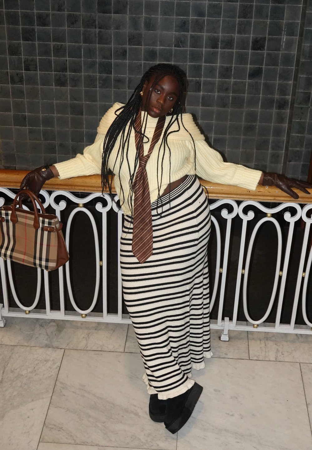 Femme aux longues tresses portant des gants marron, une jupe rayée, un pull crème et une cravate marron, appuyée sur une rambarde tenant un sac à main à carreaux.