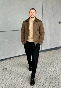 Ung mann som smiler, går på flislagt underlag, iført brun jakke, beige genser, svarte bukser og svarte sko foran en grå vegg.