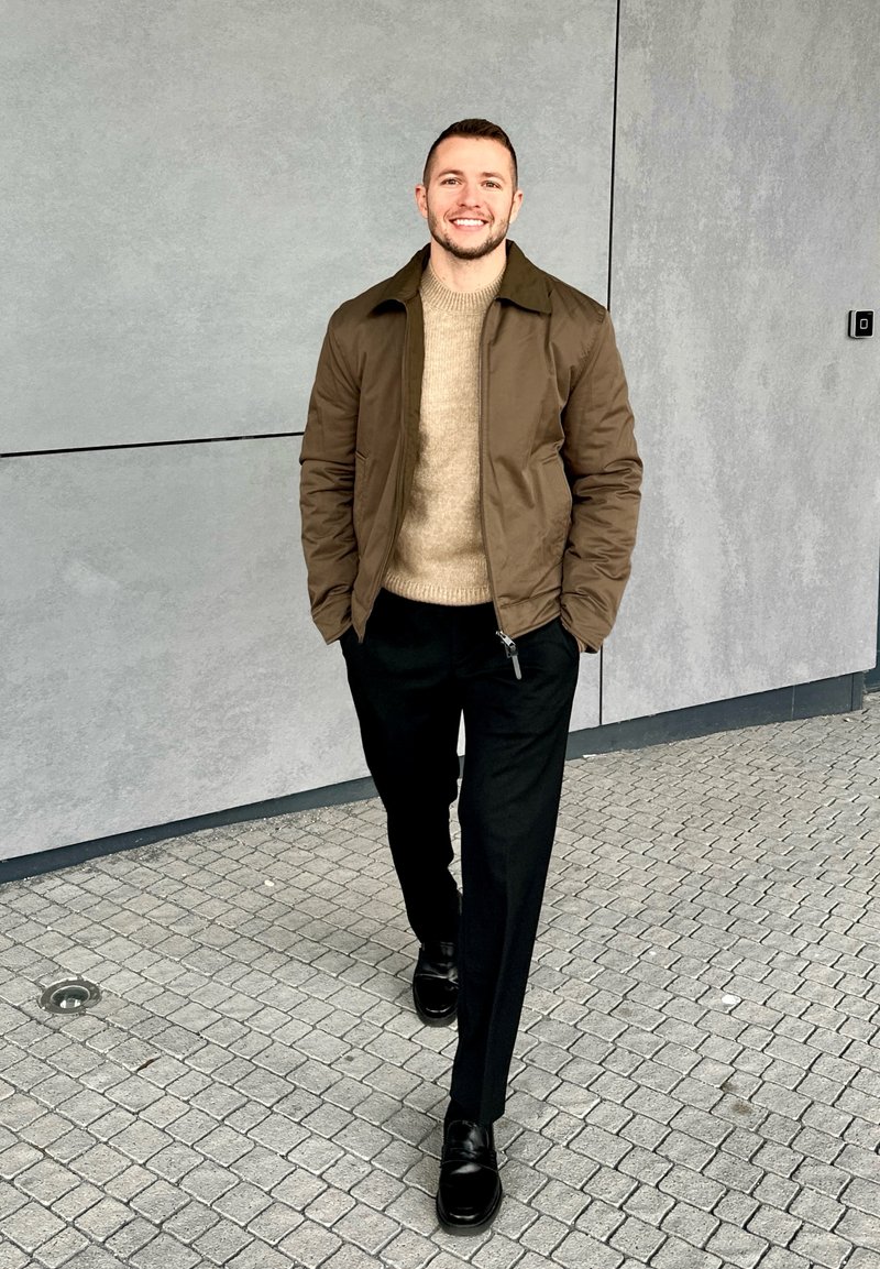 Young man smiling, walking on tiled pavement, wearing a brown jacket, beige jumper, black trousers, and black shoes against a grey wall.