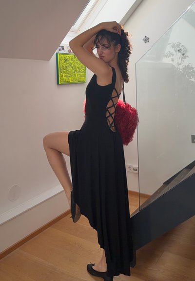 Mujer con un vestido negro de espalda abierta con cordones, posando en el interior sobre un suelo de madera junto a una escalera de cristal, sosteniendo un bolso rojo y esponjoso.