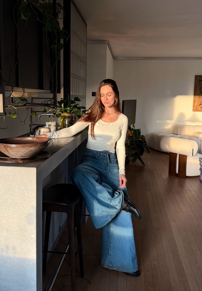 Femme aux longs cheveux portant un haut blanc et un jean à jambes larges, assise sur un tabouret dans une cuisine moderne baignée de lumière avec des plantes.