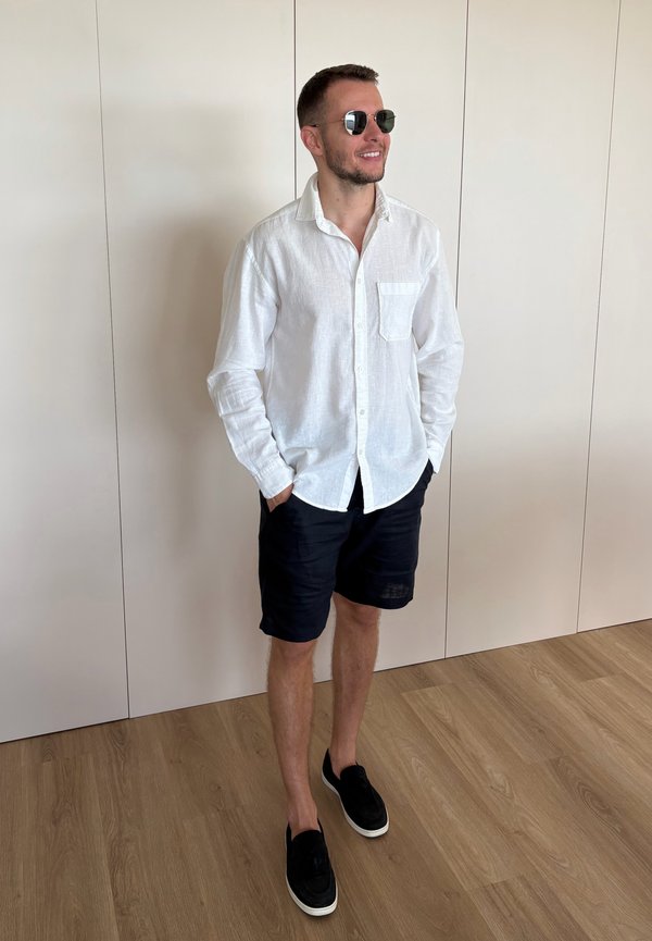 Man met zonnebril, wit shirt met lange mouwen, zwarte korte broek en zwarte instapschoenen, staand op houten vloer tegen een witte muur.