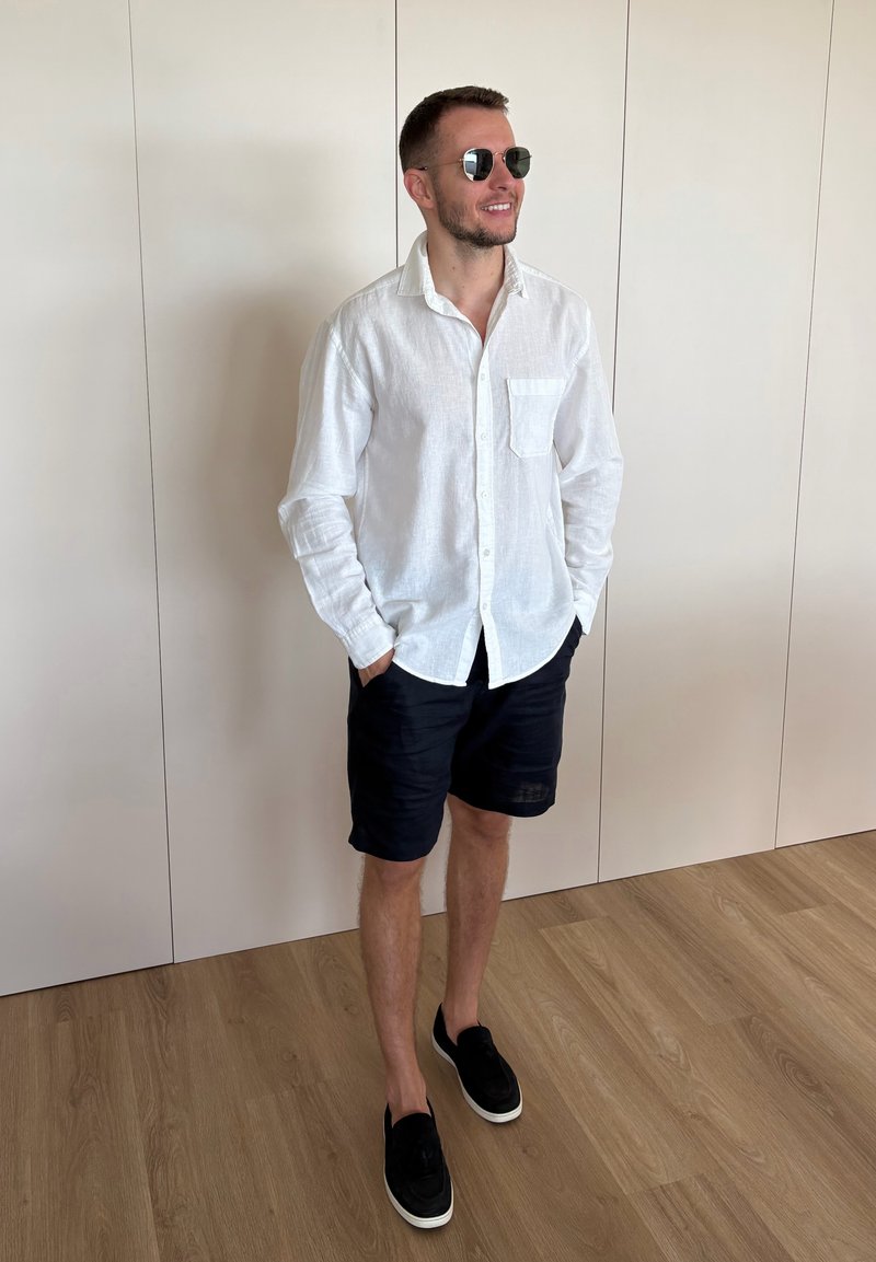 Homme portant des lunettes de soleil, une chemise blanche à manches longues, un short noir et des chaussures noires sans lacets, debout sur un sol en bois contre un mur blanc.