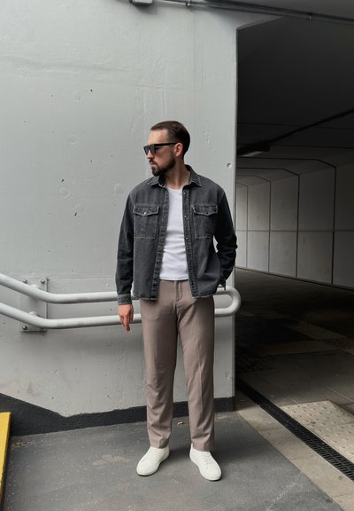 Chaqueta de mezclilla negra sobre una camiseta blanca, pantalones beiges y zapatillas blancas; de pie en un entorno urbano moderno y austero con paredes grises.