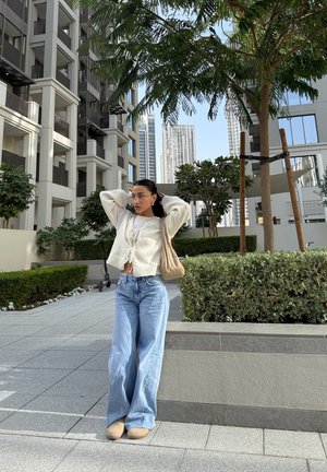 Joven mujer con suéter beige y jeans está de pie en la acera urbana cerca de árboles y edificios modernos bajo un cielo despejado.