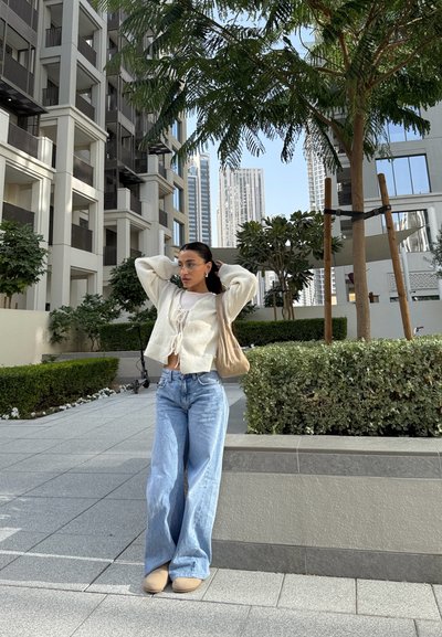 Joven mujer con suéter beige y jeans está de pie en la acera urbana cerca de árboles y edificios modernos bajo un cielo despejado.