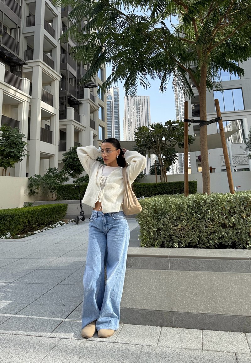 Jeune femme en pull beige et jean se tient sur un trottoir urbain près d'arbres et de bâtiments modernes sous un ciel clair.