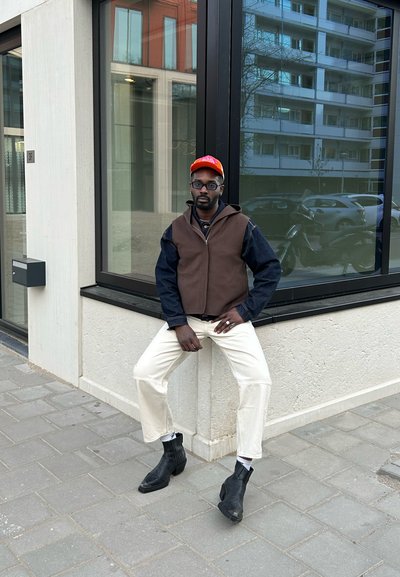 Chaleco marrón sin mangas sobre una camisa de mezclilla oscura, pantalones cortos de color blanco roto y botas negras hasta el tobillo, complementados con una gorra naranja.