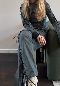 Femme aux longs cheveux blonds assise sur une chaise vert foncé, portant une veste grise à lacets et un pantalon assorti avec des bottines beige aux chevilles.