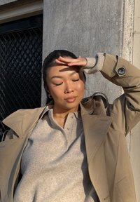 Lichtbeige trenchcoat over een grijze trui met een kraag met knopen; zachte texturen en contrasterende tinten tegen een getextureerde achtergrond.