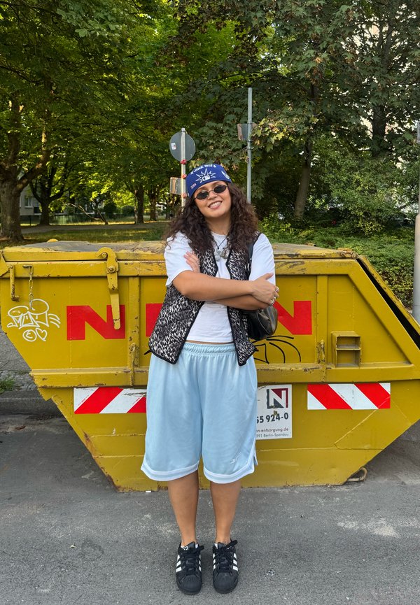 Femme portant un bandana, des lunettes de soleil, un t-shirt blanc, un gilet et un short, se tient les bras croisés devant une benne métallique jaune dans une rue bordée d’arbres.