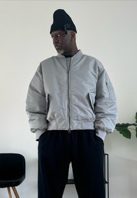 Veste bomber grise avec fermeture éclair frontale, poignets côtelés et poches latérales. Associée à un pantalon noir, sur un fond clair avec des plantes.