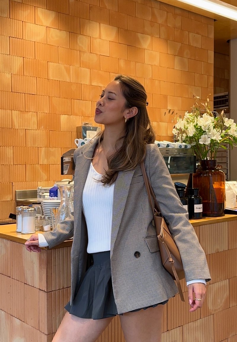 Blazer à carreaux gris sur un haut blanc côtelé, avec une mini-jupe plissée noire. Sac à main marron, cadre intérieur avec boiseries et fleurs.