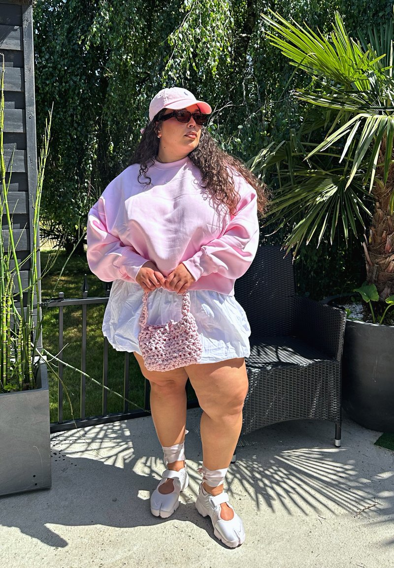 Femme portant un sweat-shirt rose, une jupe blanche, une casquette et des baskets, tenant un sac rose au crochet, debout à l'extérieur près de plantes et d'une chaise noire.
