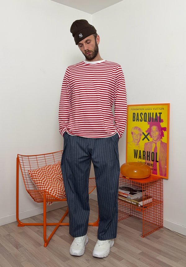 Hombre con gorro marrón, camisa roja a rayas, pantalones de rayas diplomáticas y zapatillas blancas, de pie junto a una silla de alambre naranja y un cartel de Basquiat-Warhol.