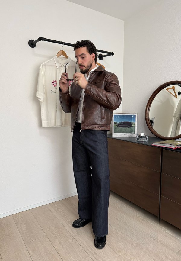 Mężczyzna w brązowej skórzanej kurtce i ciemnych dżinsach, trzymający okulary przeciwsłoneczne, stoi obok wiszącej białej haftowanej koszuli i drewnianej komody z okrągłym lustrem.