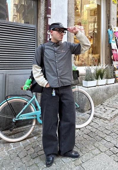 Hombre con gafas y camisa bicolor ajustando una gorra negra, de pie junto a una bicicleta azul claro en una calle empedrada con macetas y escaparate.