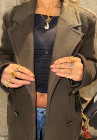 Colliers superposés en or et argent, plusieurs bagues, un manteau foncé texturé et un haut noir ajusté, mains ornées de bijoux décoratifs.