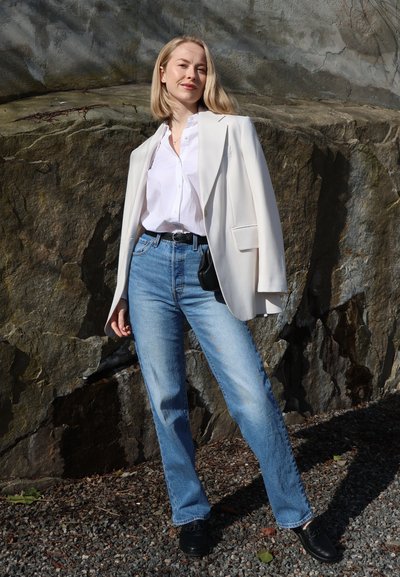 Mujer con blazer blanco colocado sobre los hombros, camisa blanca, vaqueros azules y zapatos negros, que se mantiene con confianza frente a un fondo rocoso.