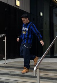 Črna in modra črtasta pulover s grafičnim besedilom, modri denim jeans in svetlo rjave škornje, ki hodi po kovinskih in kamnitih stopnicah. Podrobnost s srebrnim verigo.